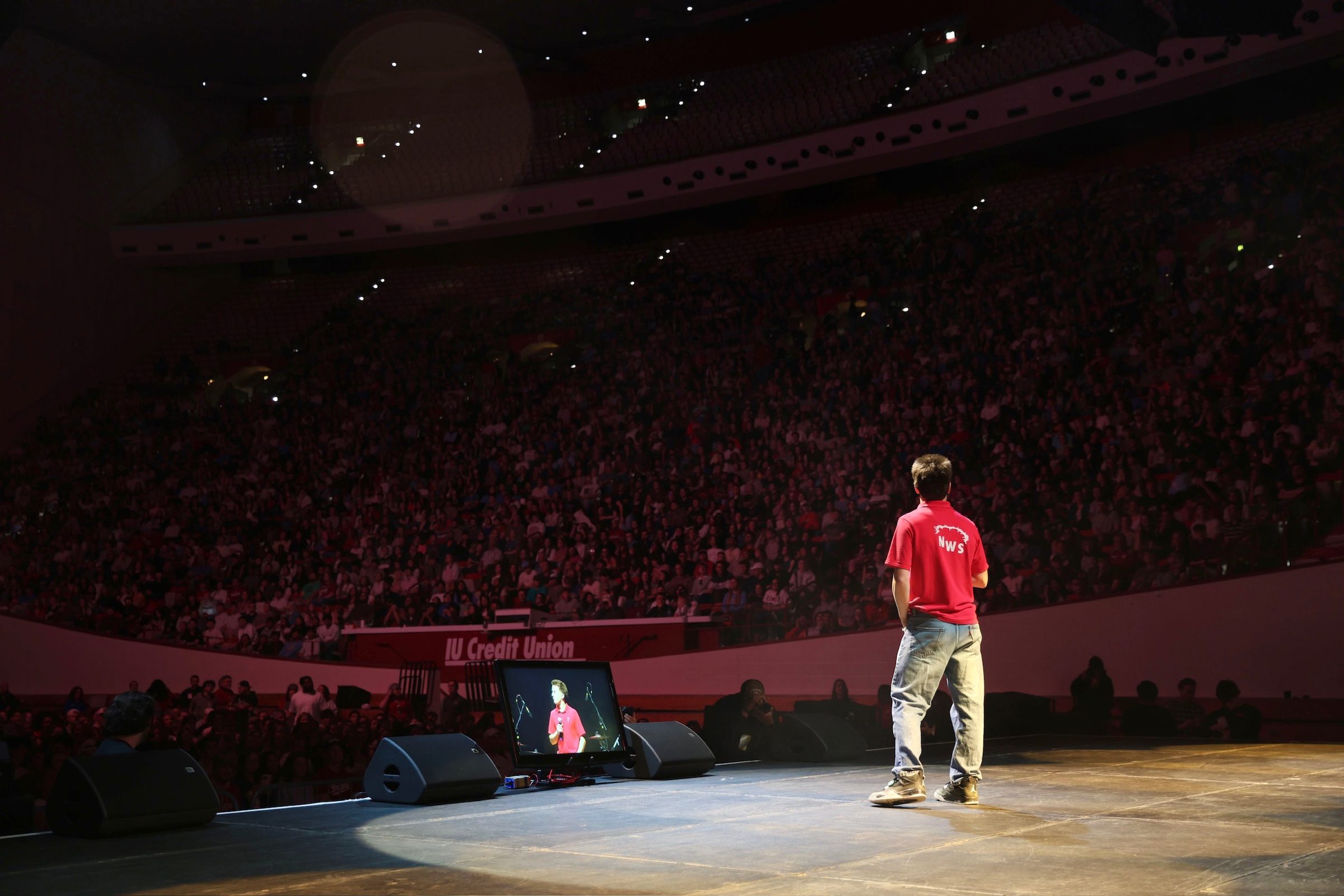 A lone figure in a red NWS polo stands at the edge of the stage facing a full Simon Skjodt Assembly Hall