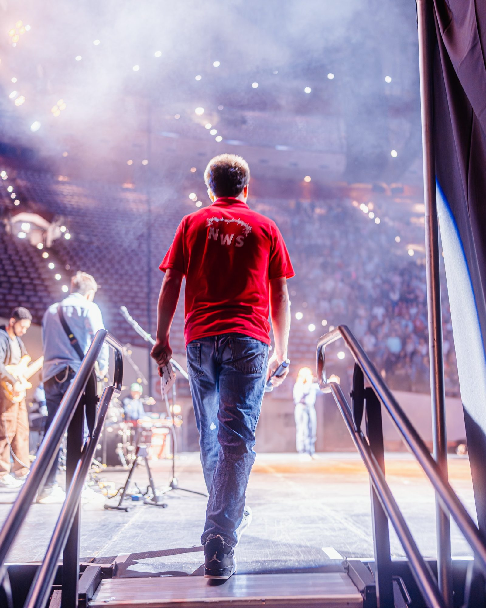 A student in a red NWS polo climbs the stage stairs toward a hazy, packed arena